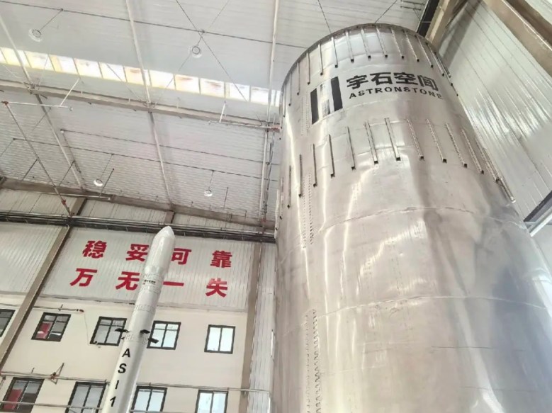 Astronstone's stainless steel rocket stage and AS-1 scale model inside a production facility in China, featuring prominent weld lines and branding; a large banner on the wall displays Chinese characters.
