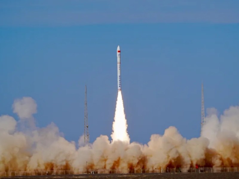 A slender, white Ceres-1 rocket with Chinese characters and a red tip ascends into a clear blue sky, leaving behind a bright trail of white-hot exhaust. Thick clouds of dust and smoke billow outward from the launch pad at Jiuquan Satellite Launch Center in the arid landscape of northwest China.