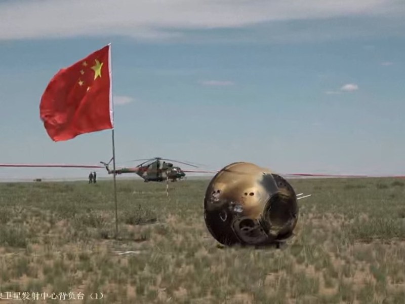 A Chinese flag stands next to a scorched, tipped over Chang'e-6 reentry capsule in the grasslands of Inner Mongolia. Helicopters are visible in the distance.