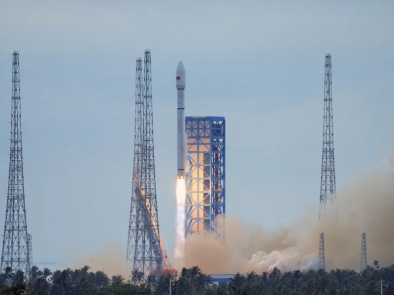 A Long March 12 rocket lifts off from the Hainan commercial launch site, rising above the pad with bright exhaust and a plume of smoke, surrounded by tall lightning towers under a pale sky.