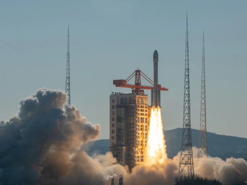 A Long March 6A rocket climbs above the launch tower at Taiyuan amid plumes of exhaust and a blue sky backdrop.