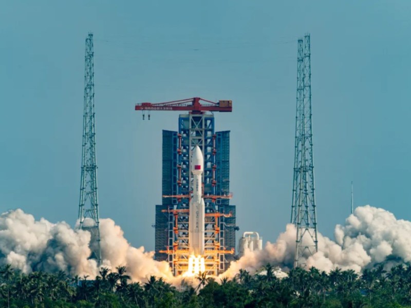 A Long March 8A rocket lifts off from Launch Pad 1 at the Hainan Commercial Space Launch Center on July 30, 2025, carrying the sixth group of Guowang LEO broadband satellites. Smoke billows around the launch pad as the vehicle ascends against a clear blue sky, framed by metal towers and palm trees in the foreground.
