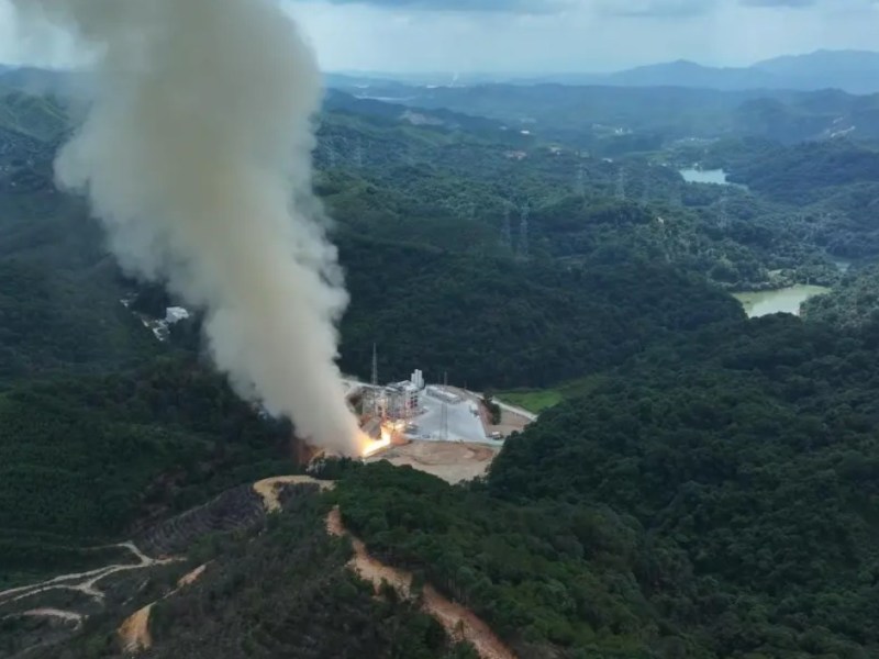 Aerial view of CAS Space's Kinetica-2 first-stage hot fire test, showing flames and thick smoke rising from a test stand nestled in forested hills.
