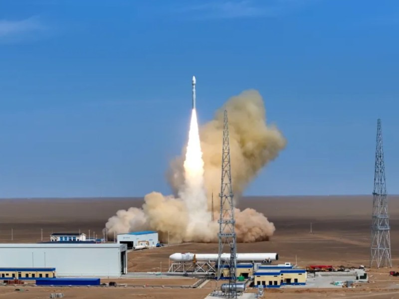 A white Kinetica-1 rocket operated by Chinese launch company CAS Space ascends into a clear blue sky, leaving behind a bright flame and thick plume of brownish exhaust. The rocket has just lifted off from a desert launch pad at the Jiuquan Satellite Launch Center in northwest China. In the foreground are low, rectangular buildings with blue roofs, a large white hangar, and tall metal towers used for communications. The barren desert landscape stretches flat into the distance under an open sky.