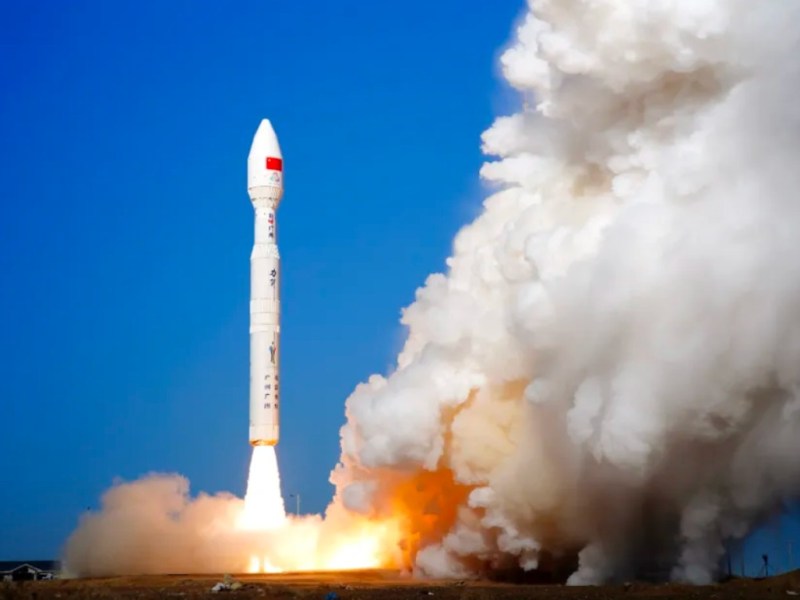 A white solid-fueled Chinese Kinetica-1 rocket lifts off vertically from a desert launch pad, producing a large plume of bright orange flame and thick white smoke against a clear blue sky.