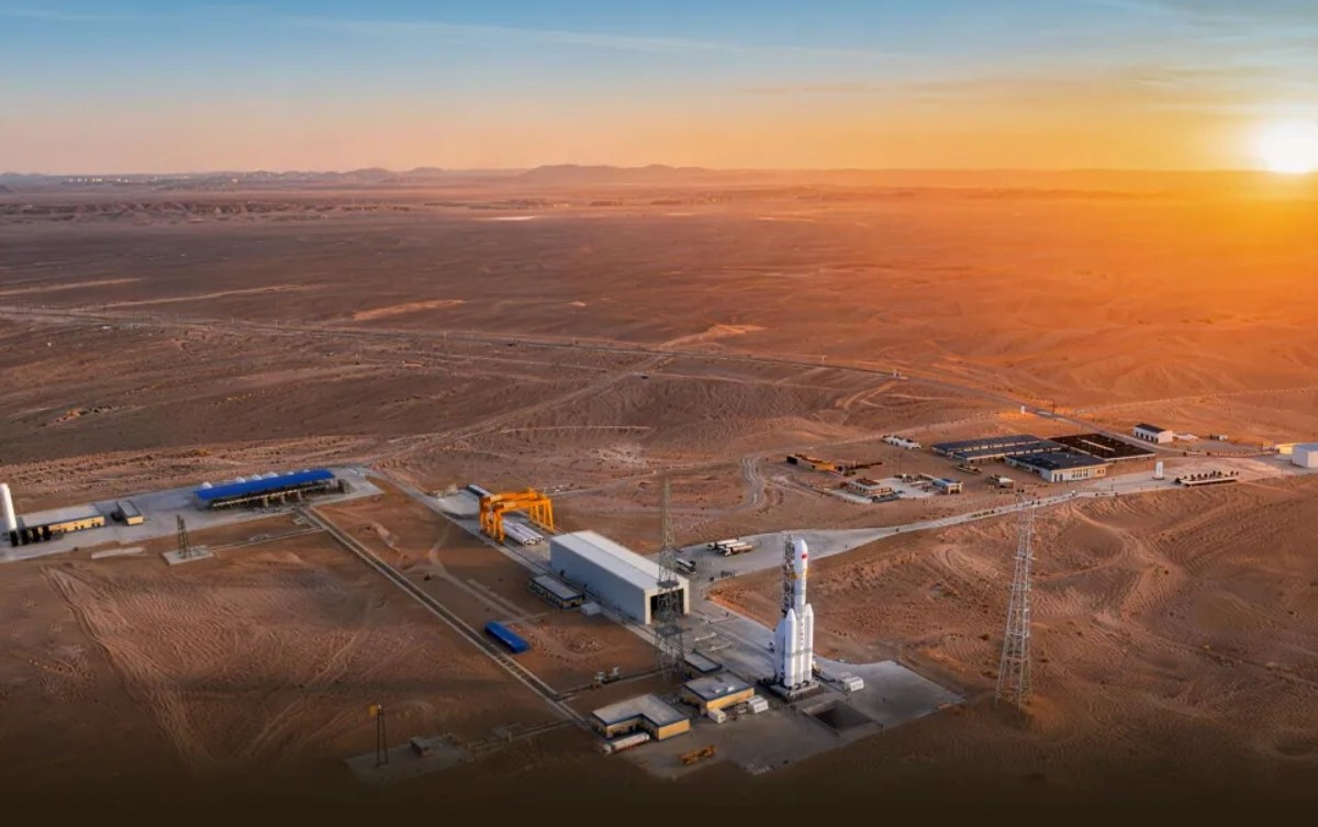 A wide aerial view of a rocket launch site in a desert landscape at sunset. A tall white rocket stands vertically on a launch pad near the center-right, surrounded by service towers and low buildings. A long rectangular assembly building and crane structure sit nearby, connected by paved roads. The surrounding terrain is flat, sandy, and sparsely developed, with distant mountains visible on the horizon under a warm orange sky.