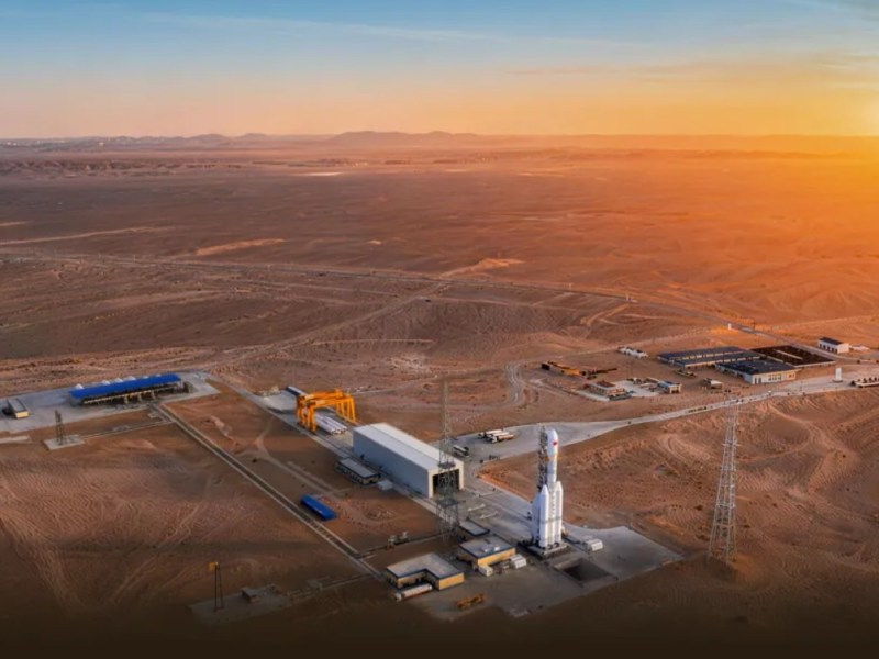 A wide aerial view of a rocket launch site in a desert landscape at sunset. A tall white rocket stands vertically on a launch pad near the center-right, surrounded by service towers and low buildings. A long rectangular assembly building and crane structure sit nearby, connected by paved roads. The surrounding terrain is flat, sandy, and sparsely developed, with distant mountains visible on the horizon under a warm orange sky.