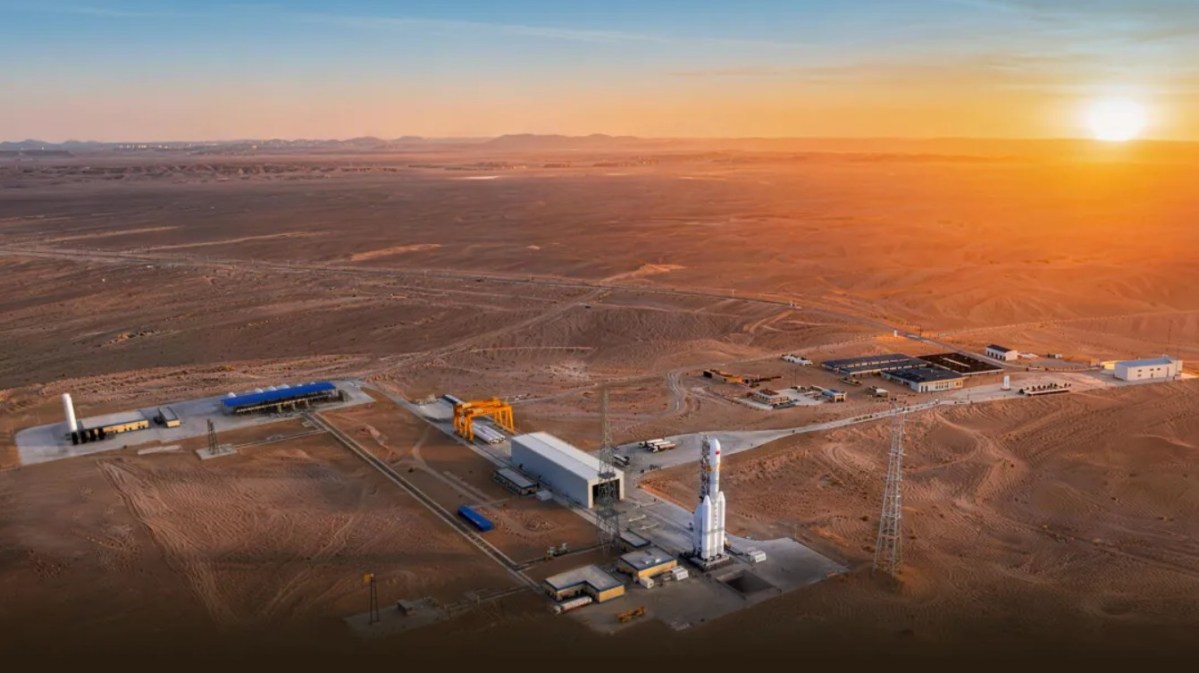 A wide aerial view of a rocket launch site in a desert landscape at sunset. A tall white rocket stands vertically on a launch pad near the center-right, surrounded by service towers and low buildings. A long rectangular assembly building and crane structure sit nearby, connected by paved roads. The surrounding terrain is flat, sandy, and sparsely developed, with distant mountains visible on the horizon under a warm orange sky.