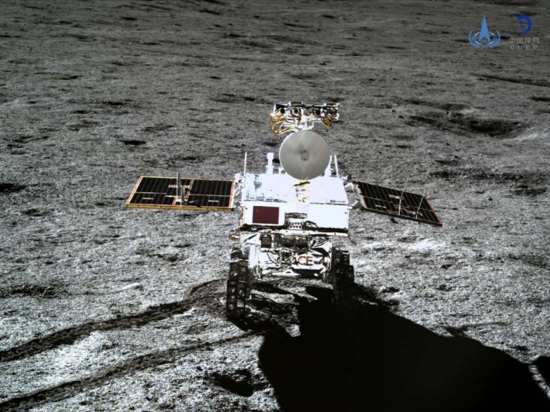 A close-up image of the Yutu-2 rover taken by the Chang’e-4 lander, showing the six-wheeled rover on the gray, uneven lunar surface of Von Kármán crater. The rover’s solar panels are extended, reflecting sunlight, with tracks visible in the regolith behind it. The barren, cratered terrain of the Moon's far side stretches into the distance.