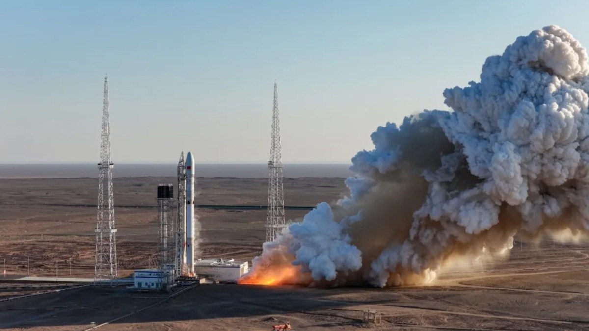A Long March rocket stands on a launch pad in a desert setting during a static fire test, with engines firing and producing a large plume of white and gray exhaust and dust spreading outward across the ground, framed by lightning towers under a clear sky.