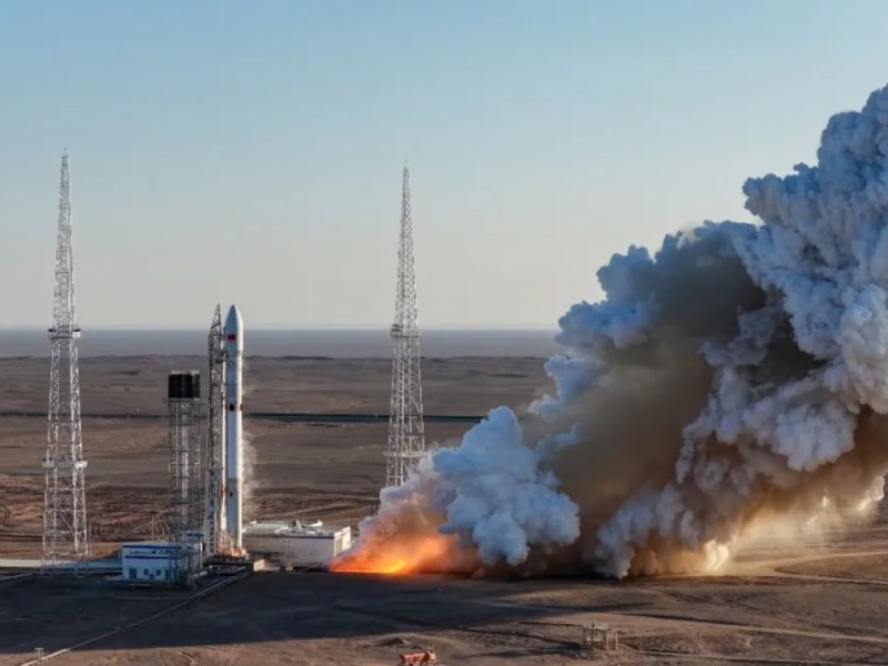 A Long March rocket stands on a launch pad in a desert setting during a static fire test, with engines firing and producing a large plume of white and gray exhaust and dust spreading outward across the ground, framed by lightning towers under a clear sky.