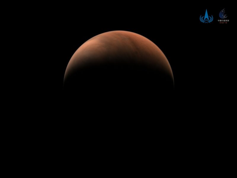 A high-resolution image of Mars taken by China's Tianwen-1 spacecraft, showing a partially illuminated view of the planet’s northern hemisphere against the darkness of space. The reddish-brown Martian surface is visible with subtle atmospheric haze.