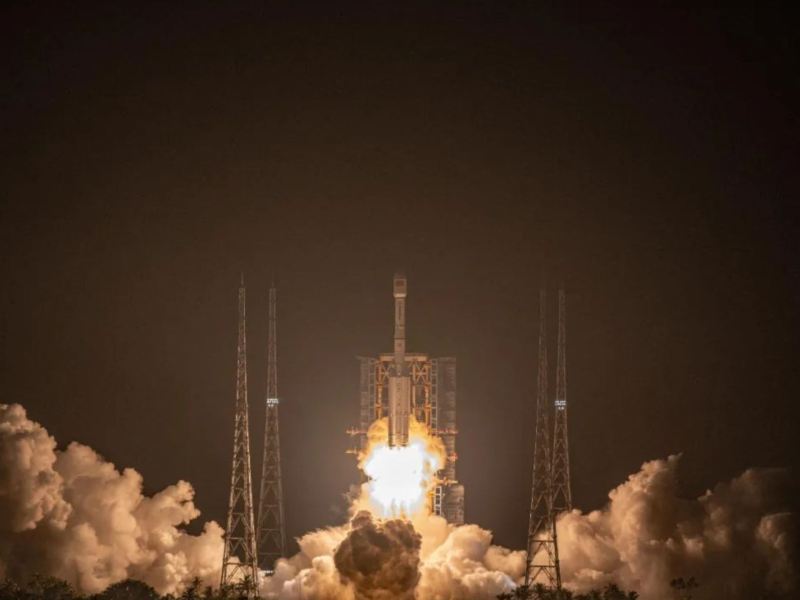 A Long March 7A rocket carrying the Zhongxing-1E satellite rises into a black sky above the launch tower at Wenchang spaceport, propelled by orange exhaust.