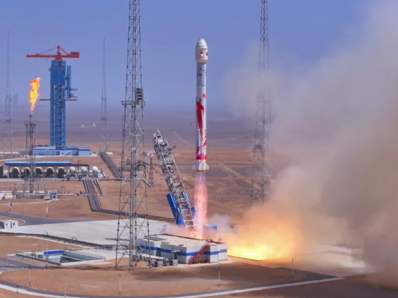 A Zhuque-2E rocket lifts off from the Dongfeng Commercial Space Launch Site in northwest China, trailing a bright exhaust plume. The rocket features red graphics on its white body. A flame stack burns off excess fuel nearby, with blue infrastructure towers and arid desert terrain visible in the background.