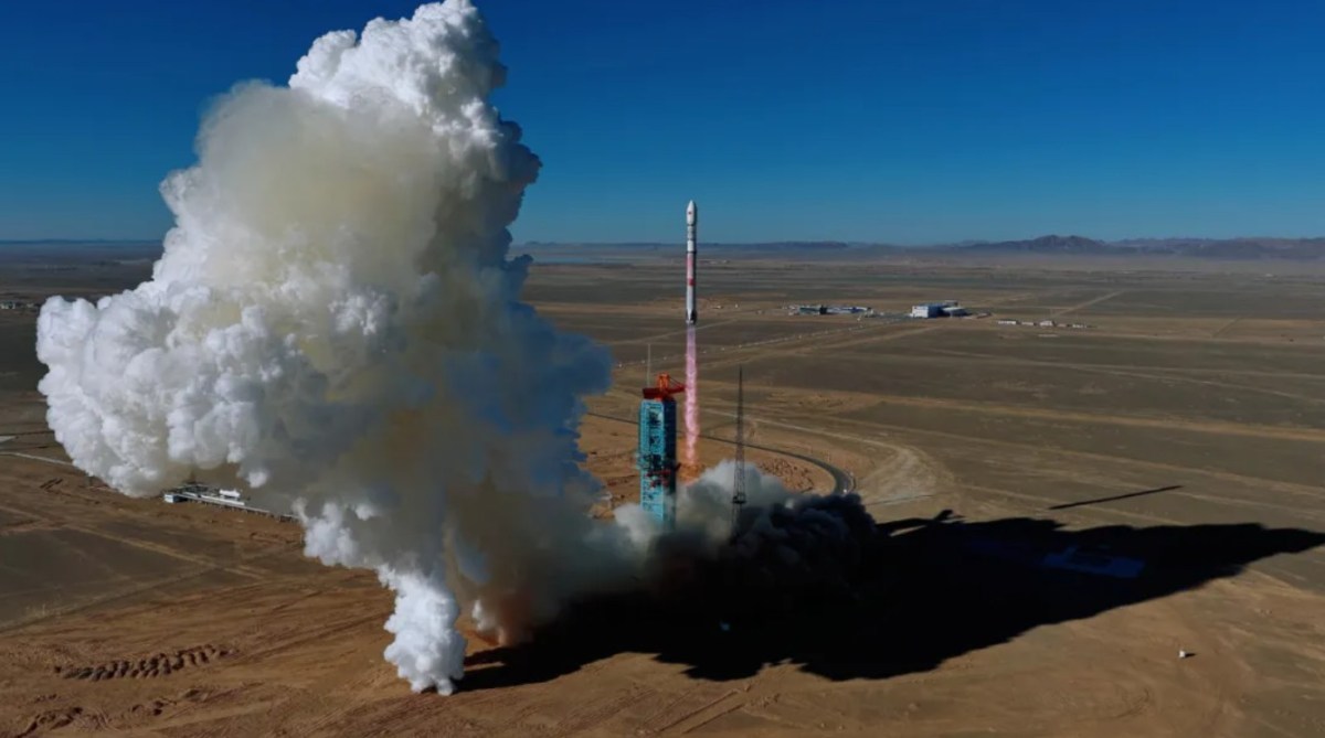 A Zhuque-3 rocket lifts off from a launch pad in a desert landscape, leaving a large plume of white exhaust behind it as it ascends into a clear blue sky. The launch tower and surrounding facilities are visible on the ground, with mountains faintly visible on the horizon.